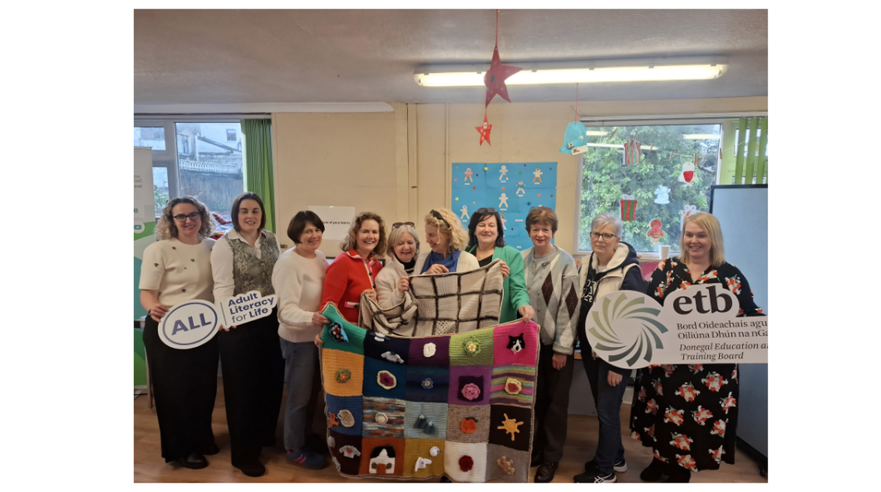 A group of women indoors holds signs for adult literacy and a colorful patchwork blanket. Decorative stars and crafts hang from the ceiling.